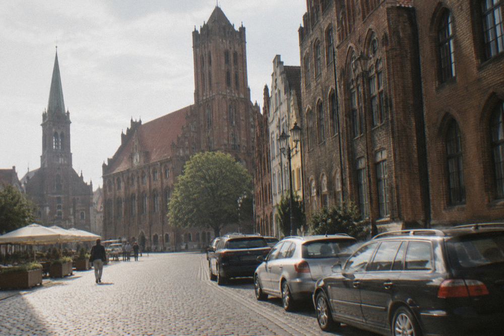 Cars parked along a cobbled street lined with Gothic brick townhouses