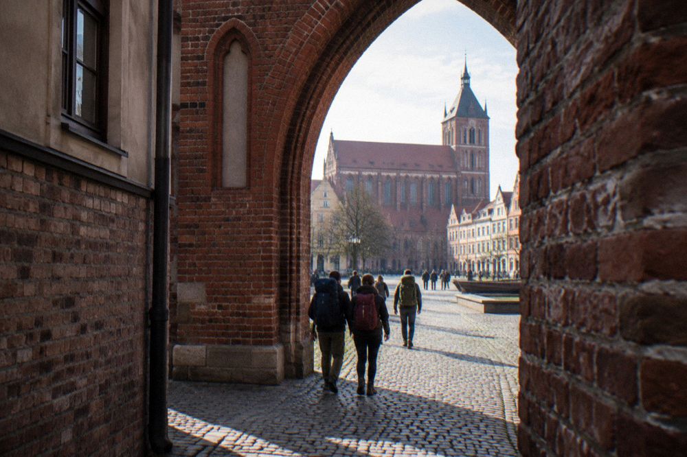 Travellers walking with backpacks through a Gothic brick gate into an old town square