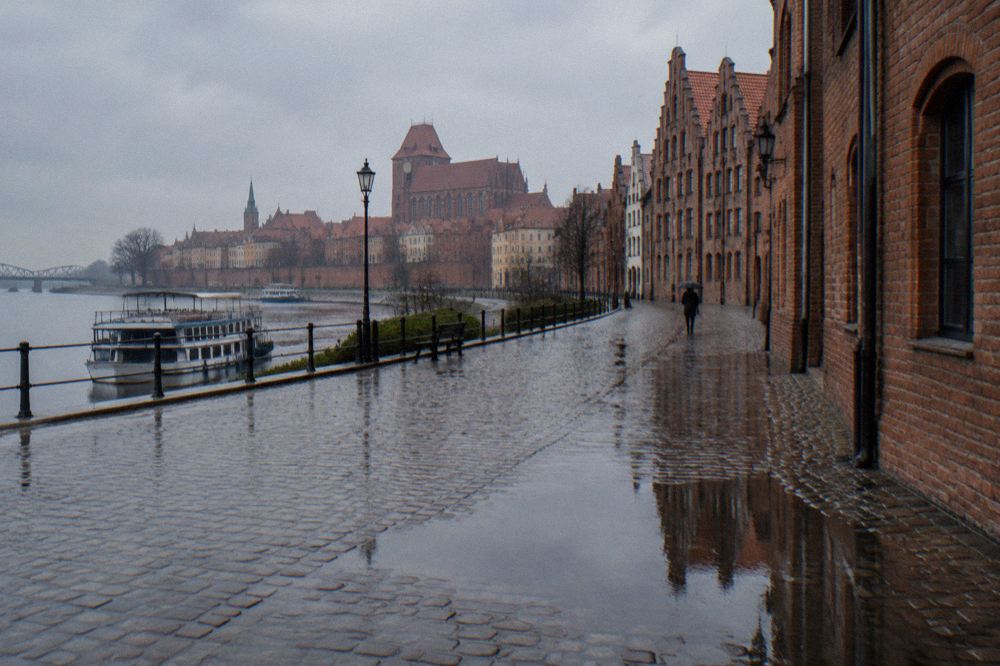 A rain-washed cobbled lane in Toruń's Old Town, wet brick gables reflected in puddles, one person with an umbrella far off.