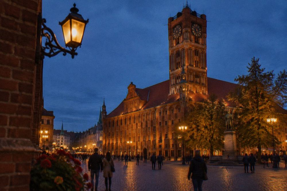 Evening view of Toruń Old Town square with lit town hall and people walking