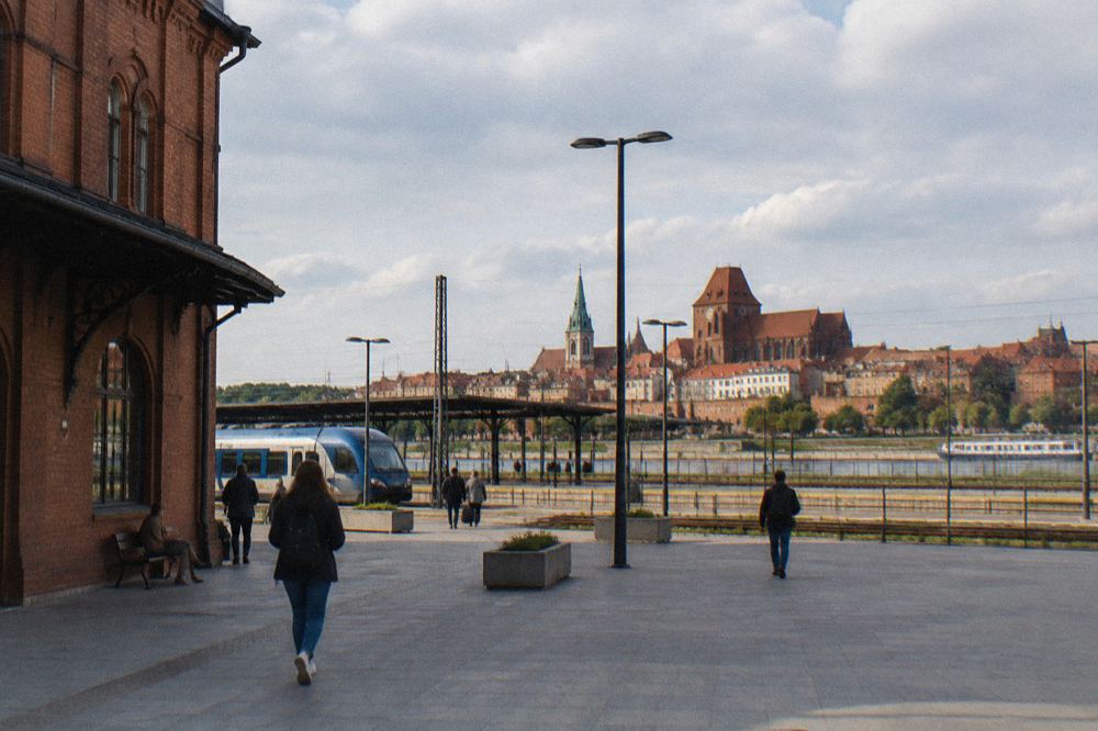 A railway station forecourt with the towers of an old town visible in the distance