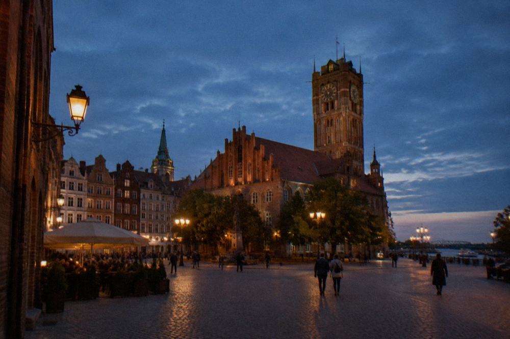 A medieval market square with gothic townhouses and a tower under an evening sky