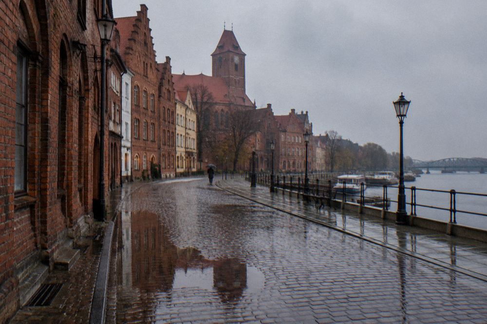 A rain-washed cobbled lane in Toruń's Old Town, wet brick gables reflected in puddles, one person with an umbrella far off.