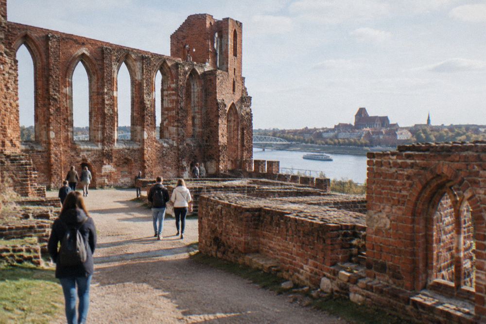 Visitors walking among the low red brick walls and arched windows of the Teutonic Castle ruins in Toruń