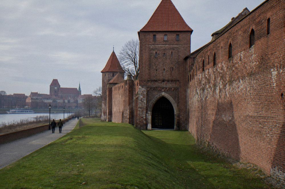 A long stretch of weathered red-brick medieval town wall with a fortified gate, grass at its foot, long shadows, Toruń.