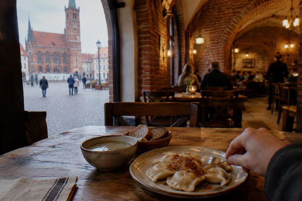 A rustic wooden table with a plate of boiled pierogi with fried onions, a bowl of soup and rye bread, a hand reaching in, in a generic brick-vaulted dining room, no writing or menu visible.