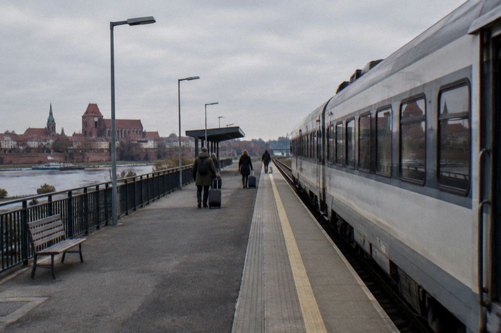 A plain unmarked regional train standing at an empty station platform in Toruń, a few travellers with suitcases small and from behind.