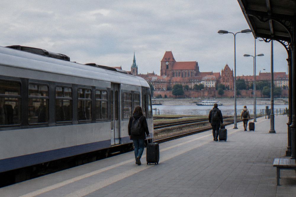 A plain unmarked regional train standing at an empty station platform in Toruń, a few travellers with suitcases small and from behind.