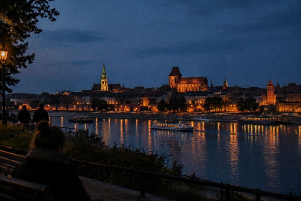 Nighttime panorama of a gothic brick old town with a river in the foreground and lit spires