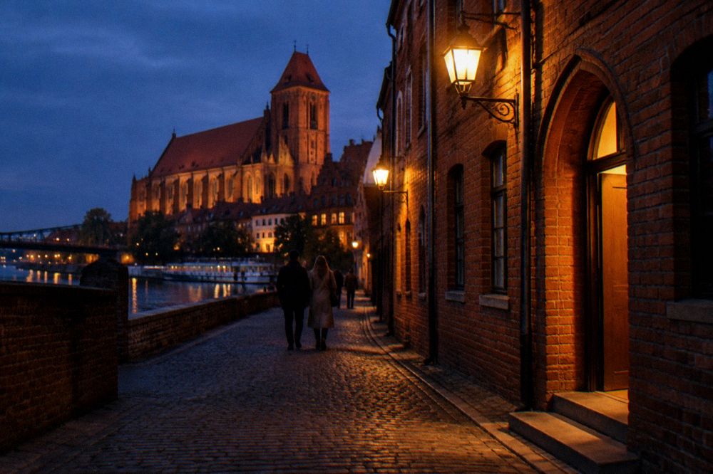 A narrow Old Town lane in Toruń after dark, warm lamps on brick walls and a lit doorway glowing, a couple walking away.