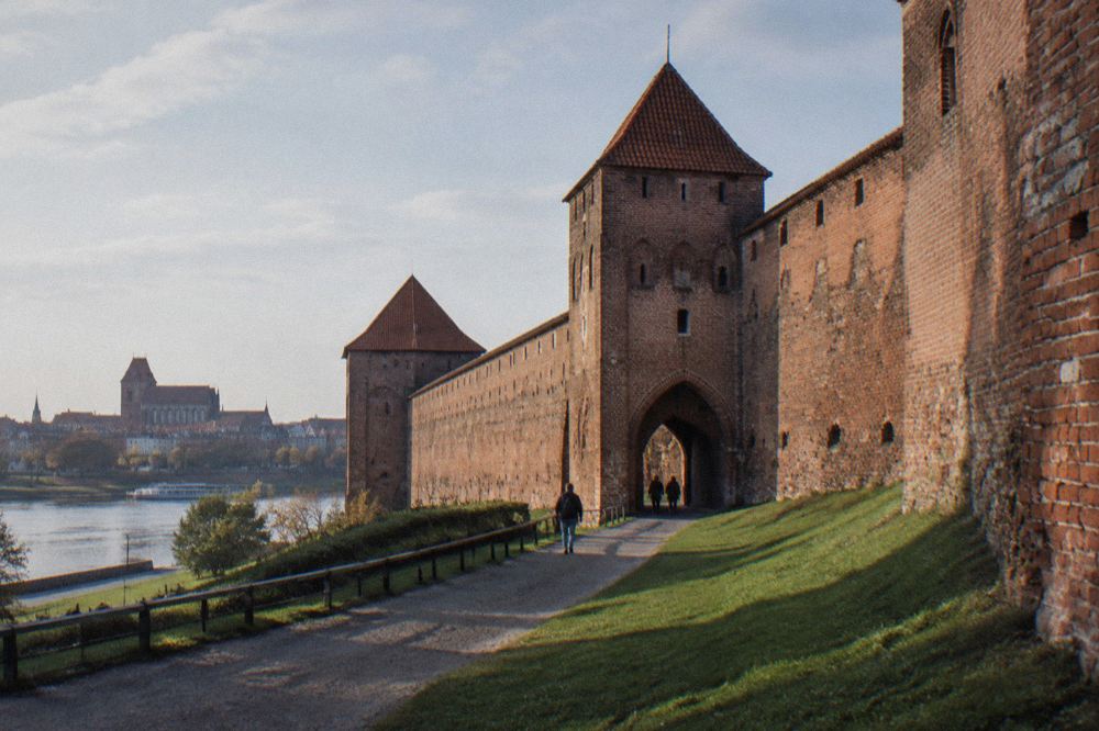 A long stretch of weathered red-brick medieval town wall with a fortified gate, grass at its foot, long shadows, Toruń.