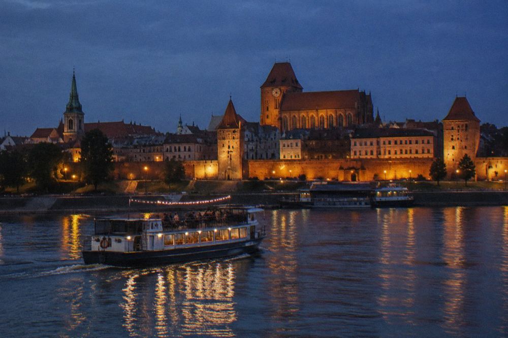 A cruise boat on the Vistula at night with the illuminated Old Town walls and towers of Toruń behind it