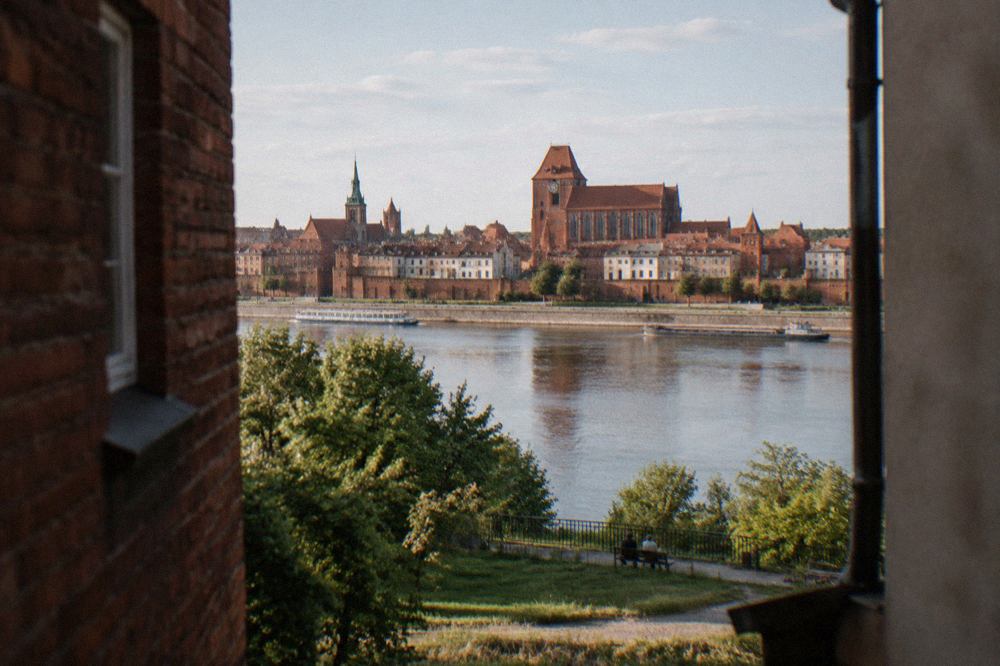 The red brick towers of Toruń Old Town seen across the Vistula river from a green viewpoint on the opposite bank