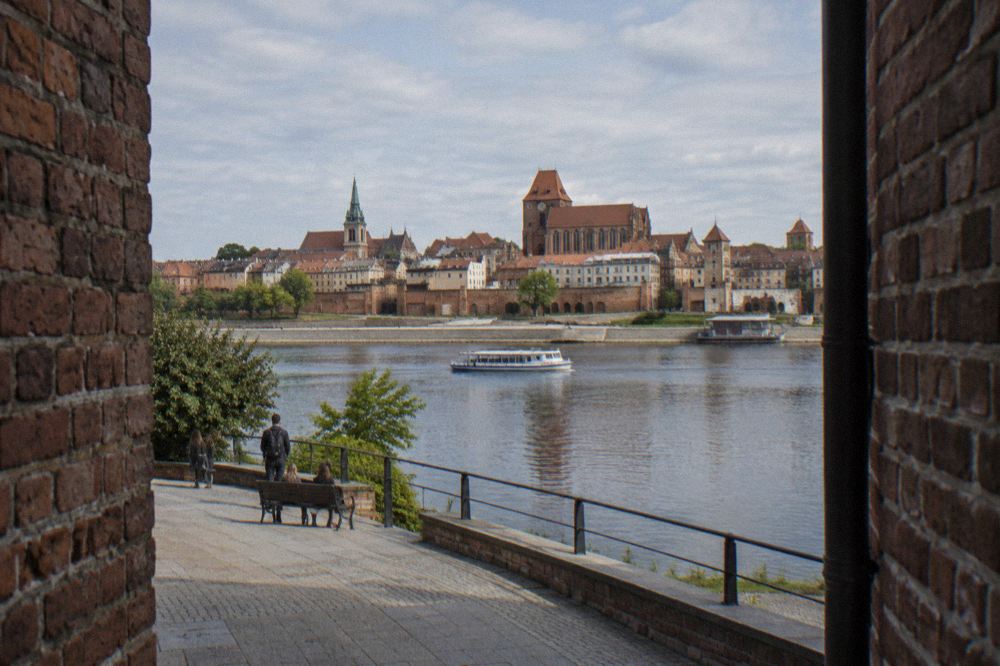 A paved viewing terrace on the left bank of the Vistula with the walled Old Town of Torun and its church towers across the water