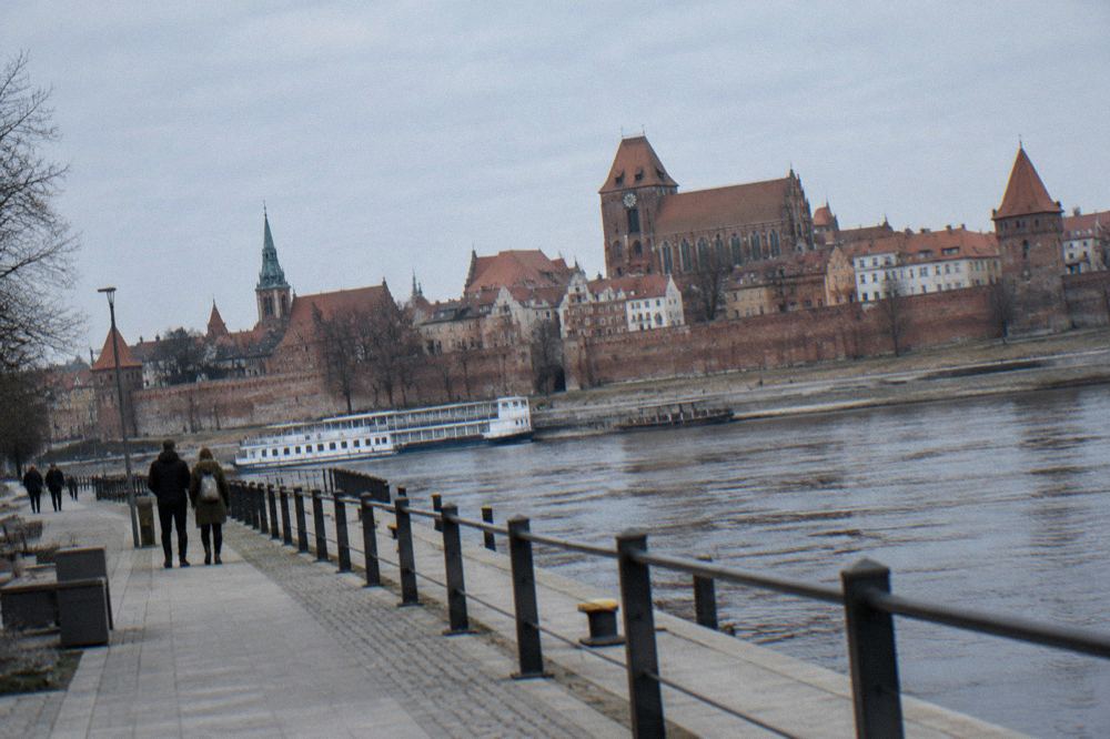 A riverside promenade along the Vistula with the brick town walls and towers of Torun rising behind it on a clear day