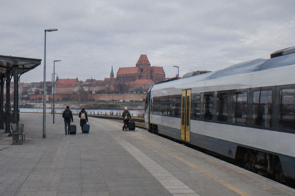 A plain unmarked regional train standing at an empty station platform in Toruń, a few travellers with suitcases small and from behind.