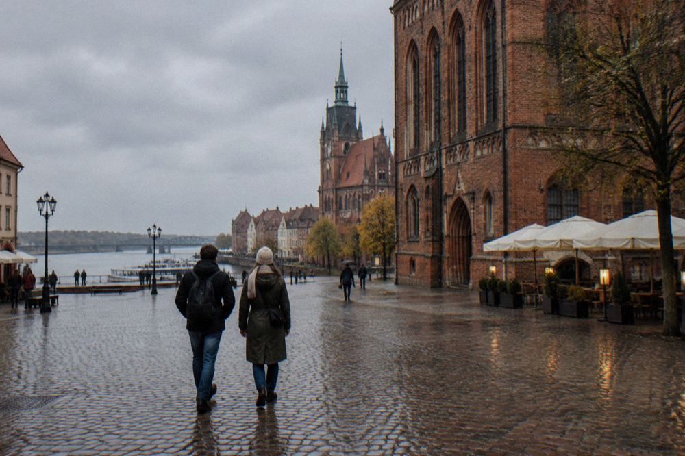 Two visitors seen from behind strolling across the cobbles of Toruń's Old Town beside a red-brick Gothic facade, cafe umbrellas at the edge.