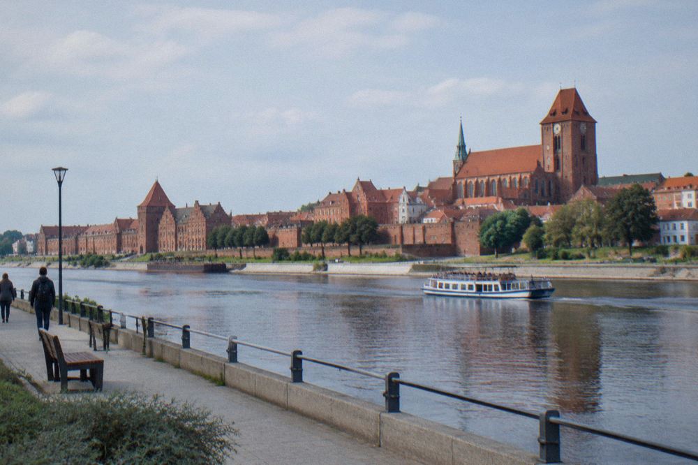 Red-brick medieval buildings and a church tower seen from a riverside boulevard