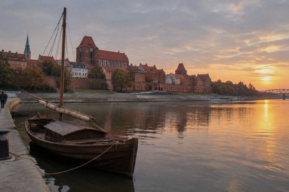 A traditional wooden boat moored on the Vistula river at golden hour below the Torun old town walls