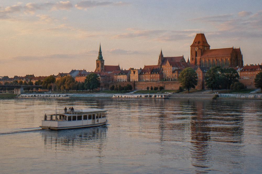 A small plain unmarked tour boat on the wide Vistula with the red-brick Toruń skyline behind, calm water, a few passengers small on deck.
