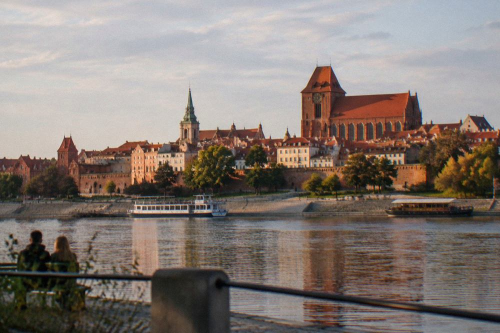 A red-brick church tower and old town rooftops of Toruń seen from across the Vistula river