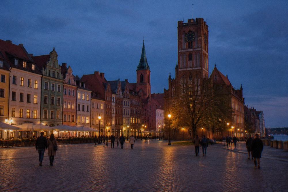 Passengers walking across a market square lined with colourful medieval townhouses at dusk