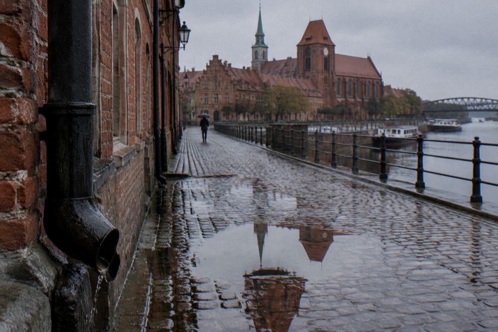 A rain-washed cobbled lane in Toruń's Old Town, wet brick gables reflected in puddles, one person with an umbrella far off.