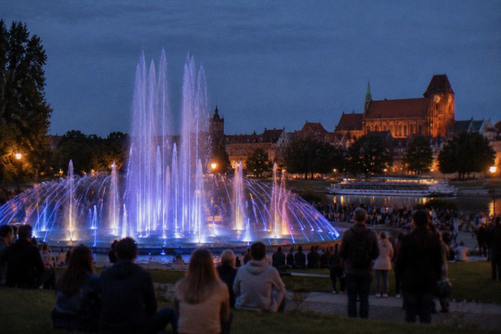 Illuminated multimedia fountain spraying water in front of crowds at dusk in a Toruń park