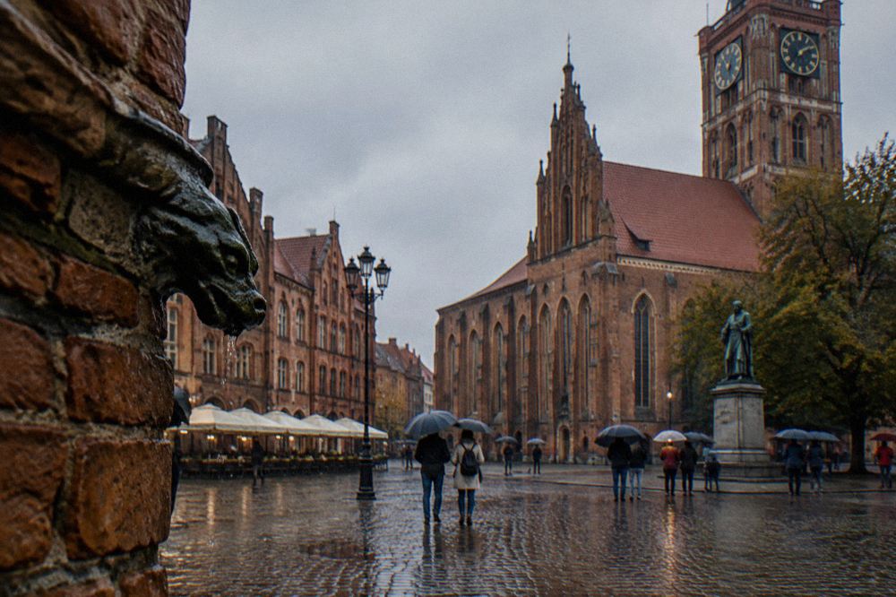 Visitors standing under umbrellas on a medieval square surrounded by brick Gothic buildings in Toruń