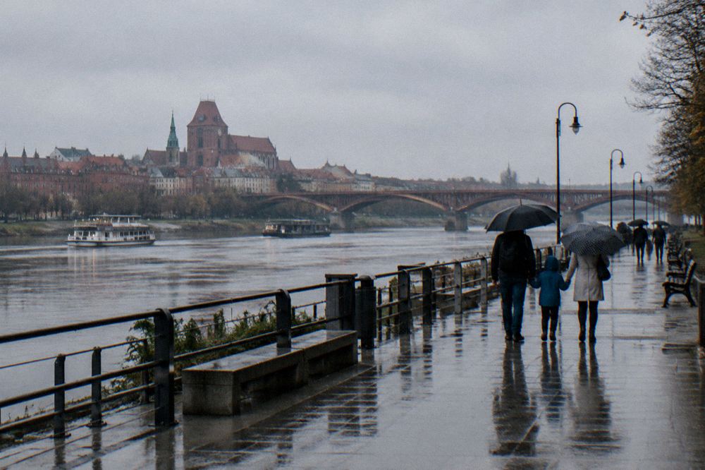 A family walking with umbrellas along a riverside boulevard with a brick bridge in the background