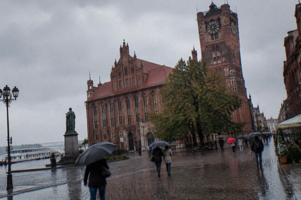 Rainy day visitors under umbrellas crossing the main square of the Toruń Old Town past the Gothic town hall